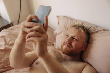 Übergewichtiger Mann, der mit seinem Smartphone ein Selfie im Bett macht, ohne ein Hemd zu tragen - MASF35036