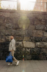 Seitenansicht eines Mannes, der an einem sonnigen Tag auf dem Fußweg vor einer Mauer geht - MASF35039