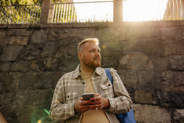 Mann hält Smartphone und schaut weg, während er an einem sonnigen Tag vor einer Wand steht - MASF35042