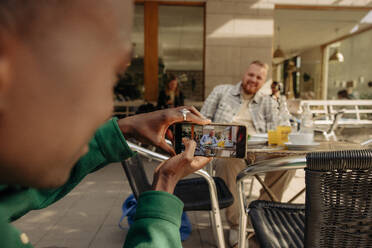 Junger Mann fotografiert männlichen Freund mit seinem Smartphone in einem Straßencafé - MASF35045