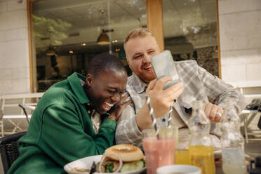 Lächelnder Mann, der sein Smartphone mit einem glücklichen männlichen Freund teilt, während er in einem Straßencafé sitzt - MASF35051