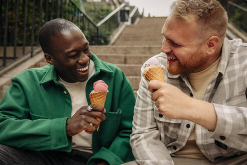 Glückliche multirassische männliche Freunde, die auf einer Treppe sitzend ein Eis genießen - MASF35059