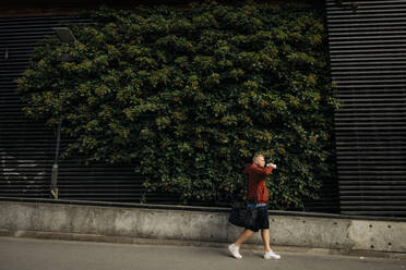 Seitenansicht eines Mannes, der Wasser trinkt, während er auf dem Fußweg an einer mit Pflanzen bewachsenen Mauer entlang geht - MASF35062