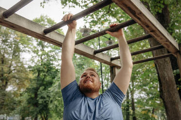 Smiling overweight man practicing monkey bars at park - MASF35066