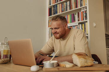 Lächelnder fettleibiger Mann, der einen Laptop benutzt, während er zu Hause auf dem Tisch sitzt und frühstückt - MASF35078