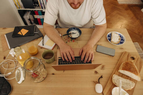 Hoher Blickwinkel des Mannes, der auf dem Laptop tippt, während er zu Hause mit dem Frühstück auf dem Tisch sitzt - MASF35079