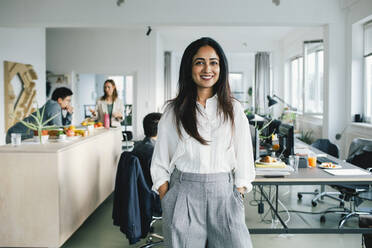 Portrait of confident smiling businesswoman standing with hands in pockets at office - MASF35109