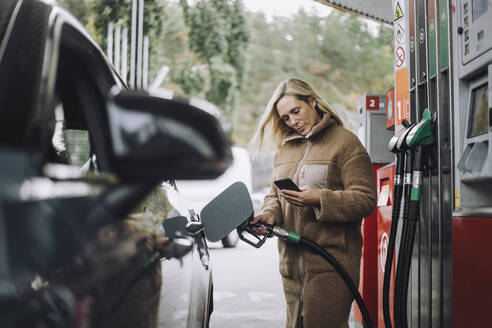 Frau benutzt Mobiltelefon beim Tanken an einer Tankstelle - MASF35156