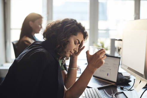 Tired businesswoman with head in hand holding drinking glass at office - MASF35223