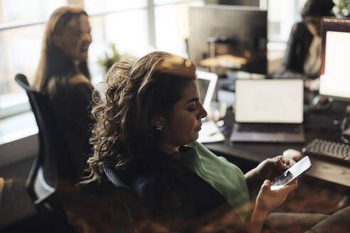 Seitenansicht einer Unternehmerin, die mit ihrem Smartphone Textnachrichten verschickt, gesehen durch ein Glas im Büro - MASF35227