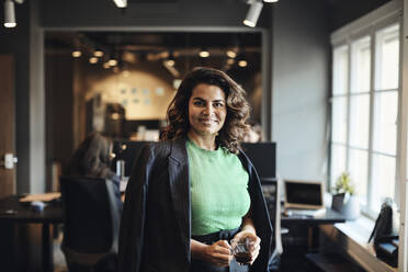 Porträt einer glücklichen Unternehmerin mit Teetasse im Büro stehend - MASF35235