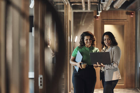 Reife Unternehmerinnen, die über einen Laptop diskutieren, während sie im Büro auf dem Flur stehen - MASF35245