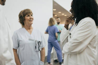 Lächelnde Ärztin mit Händen in den Taschen im Gespräch mit einem Kollegen im Krankenhaus - MASF35282