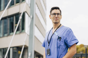 Selbstbewusster männlicher Arzt in blauem Kittel, der vor einem Krankenhausgebäude wegschaut - MASF35299