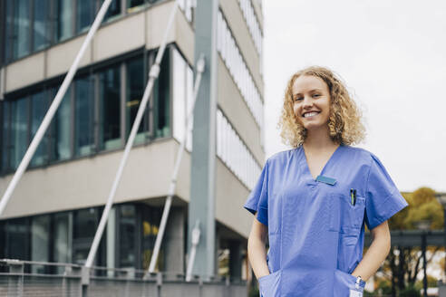 Porträt einer glücklichen Krankenschwester mit Händen in den Taschen vor einem Krankenhaus stehend - MASF35300