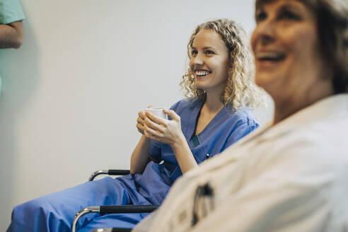 Glückliche Krankenschwester, die eine Kaffeetasse hält, während sie auf einem Stuhl neben einem Kollegen sitzt - MASF35317