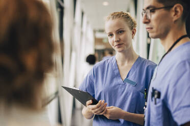 Junges weibliches Krankenhauspersonal hält ein Klemmbrett, während es mit Kollegen diskutiert - MASF35351