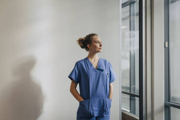 Nachdenkliche junge Krankenschwester steht mit den Händen in den Taschen an der Wand eines Krankenhauses - MASF35371