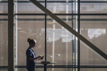 Seitenansicht einer Krankenschwester, die einen Tablet-PC benutzt, während sie am Fenster eines Krankenhauses steht - MASF35377