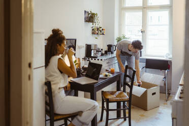 Mann beim Auspacken eines Kartons in der Küche mit seiner Freundin, die zu Hause auf dem Esstisch sitzt - MASF35390