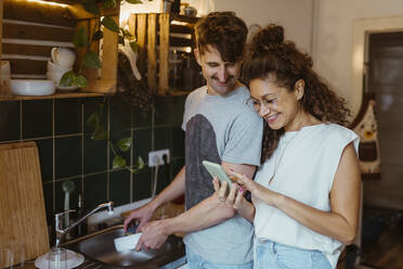 Smiling man doing dishes while looking at smart phone held by girlfriend in kitchen - MASF35391