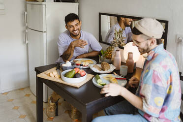Glückliches homosexuelles Paar, das Früchte isst, während es am Esstisch in der Küche sitzt - MASF35414