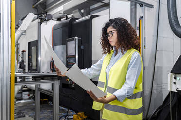 Young engineer examining blueprints standing by machine in industry - AAZF00040