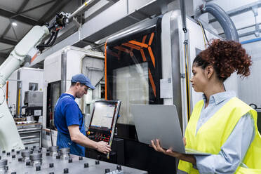 Engineer holding laptop and standing by colleague in industry - AAZF00056