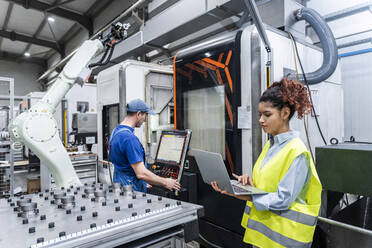 Young engineer using laptop with colleague operating machine in factory - AAZF00057