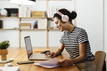 Smiling freelancer wearing wireless headphones working on laptop at table - EBSF02746