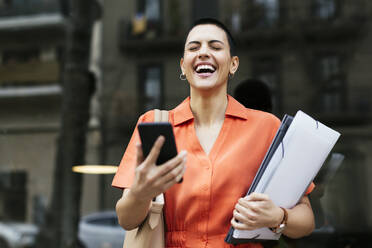 Geschäftsfrau mit Mobiltelefon und Akten lachend vor einer Glaswand - EBSF02759
