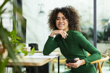 Happy businesswoman with curly hair holding mobile phone at table - EBSF02763