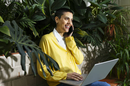 Glückliche Geschäftsfrau im Gespräch über Smartphone mit Laptop vor Pflanzen sitzend - EBSF02766
