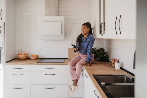Smiling woman sitting on kitchen counter with smart phone at home - EBBF07942