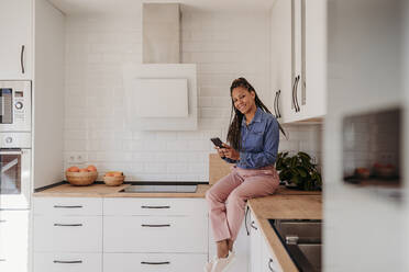 Lächelnde reife Frau sitzt auf Küchentisch mit Smartphone zu Hause - EBBF07943