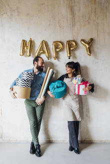 Happy couple holding gift boxes leaning on wall - MEUF08891