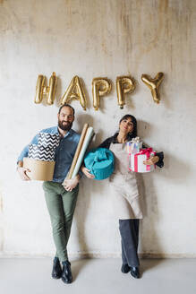 Smiling couple holding gift boxes leaning on wall - MEUF08892