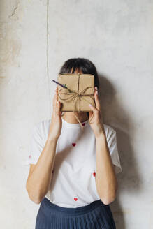 Frau bedeckt ihr Gesicht mit einem Geschenkkarton vor einer Wand - MEUF08904