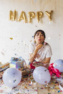 Woman blowing confetti standing in front of wall - MEUF08920