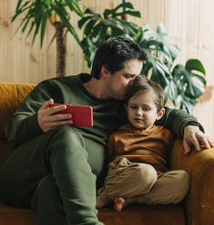 Father sitting with smart phone and kissing son at home - VSNF00424