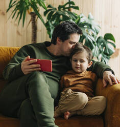 Vater sitzt mit Smartphone und küsst Sohn zu Hause - VSNF00424