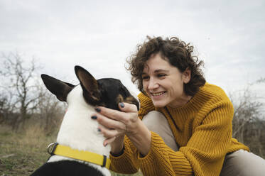 Happy woman wearing sweater petting dog - ALKF00101