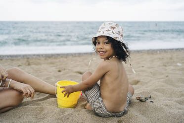 Happy cute boy wearing hat playing with bucket in sand - JJF00048