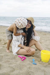 Boy kissing mother sitting on sand at beach - JJF00051