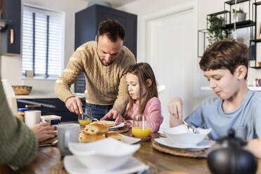 Vater schmiert Butter auf Brot, während die Familie in der Küche frühstückt - WPEF07210