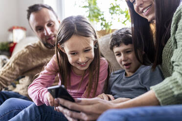 Happy girl using smart phone with family sitting on sofa at home - WPEF07248