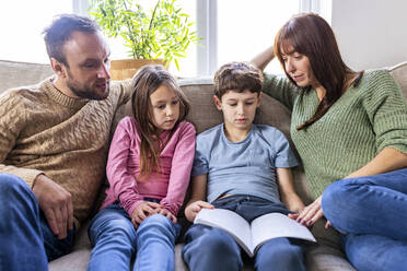 Boy reading book with family on sofa in living room - WPEF07252