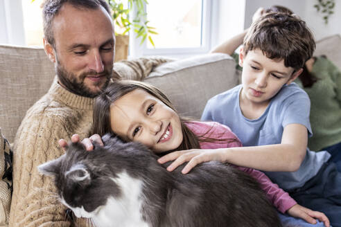 Glücklicher Vater mit Tochter und Sohn streichelt Katze auf dem Sofa zu Hause - WPEF07255
