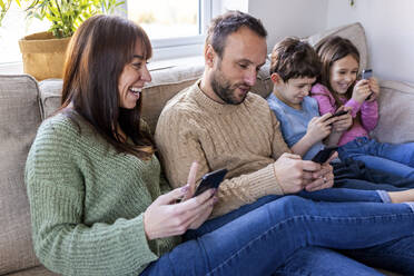 Happy woman with family using mobile phones on sofa at home - WPEF07264