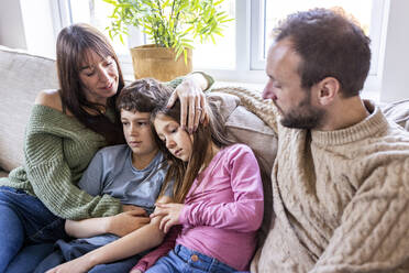 Glückliche Frau, die ihre Kinder umarmt, mit einem Mann, der zu Hause auf dem Sofa sitzt - WPEF07270